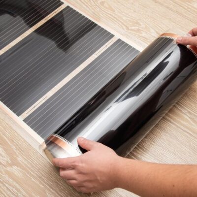 Manos desenrollando una lámina para calefacción por suelo radiante sobre un piso de madera clara.